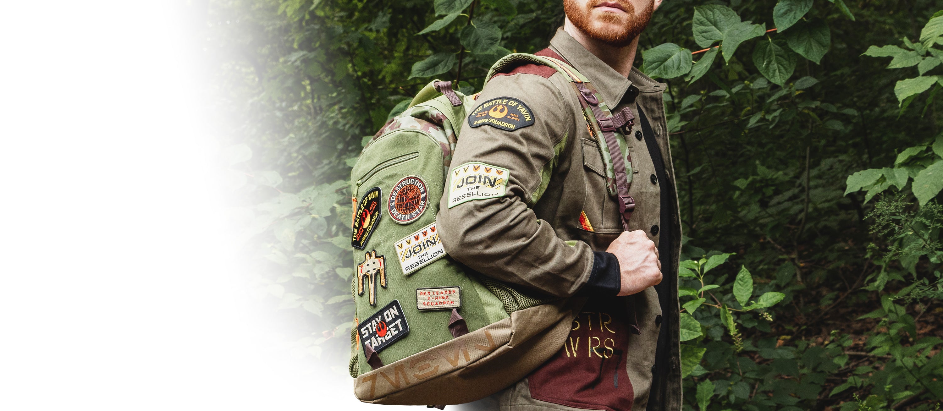 Person wearing a military-style jacket with patches and a backpack, standing in front of green foliage.