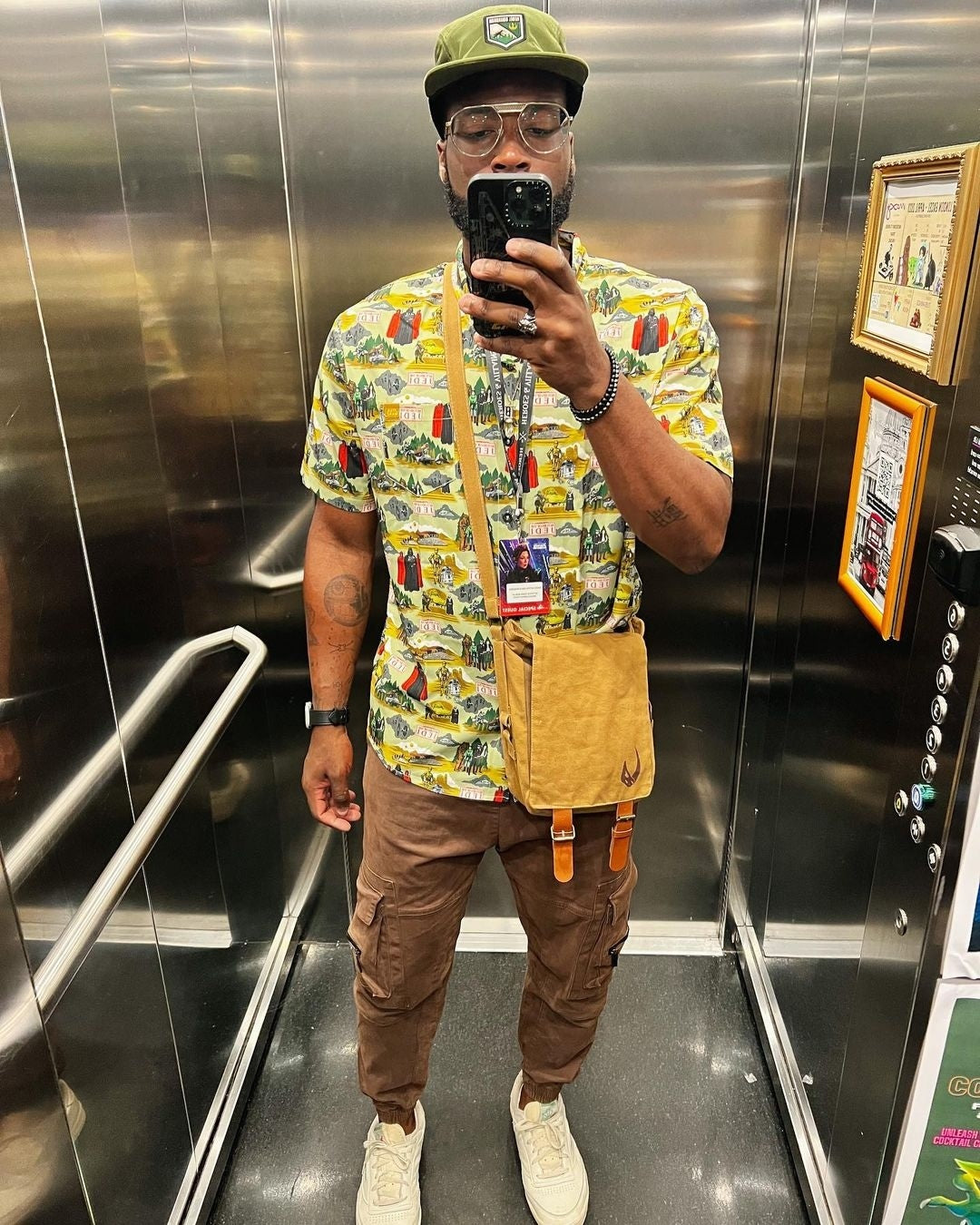 Person taking a mirror selfie in an elevator wearing a colorful shirt and brown pants.
