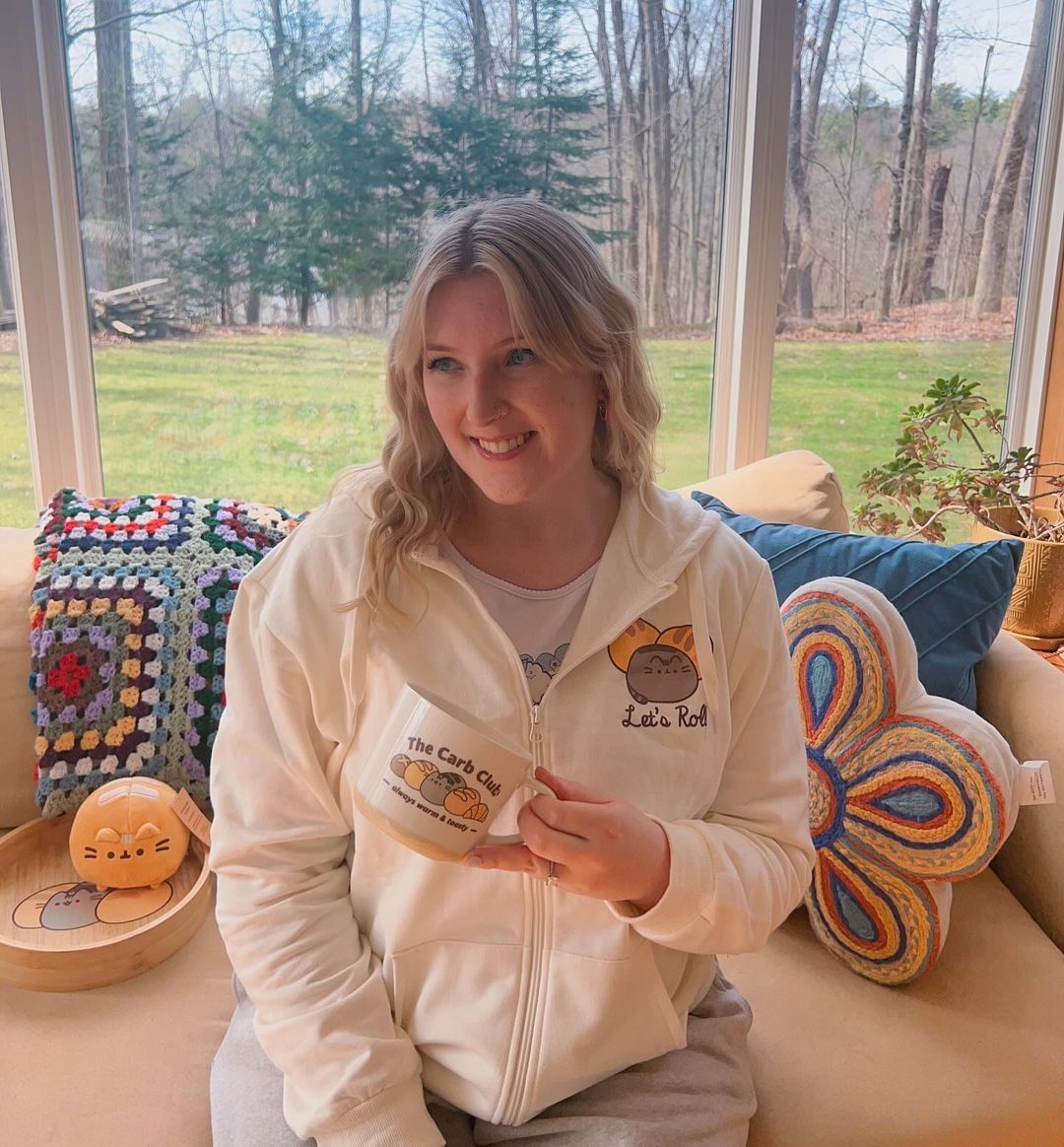 Person wearing a cream-colored hoodie with a logo, sitting on a couch in a room with large windows showing a garden view.