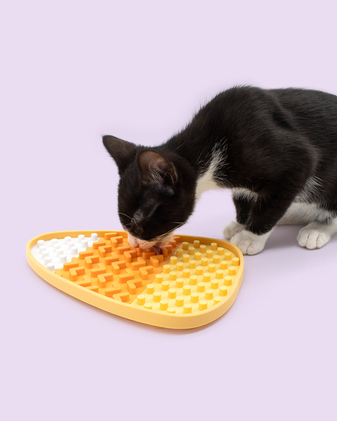A black and white cat interacting with a yellow, orange, and white textured silicone puzzle mat on a solid light purple background.