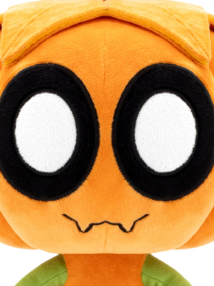 Close-up of a plush toy face in bright orange with oversized black and white eyes and a small black stitched mouth, with green fabric visible at the bottom.
