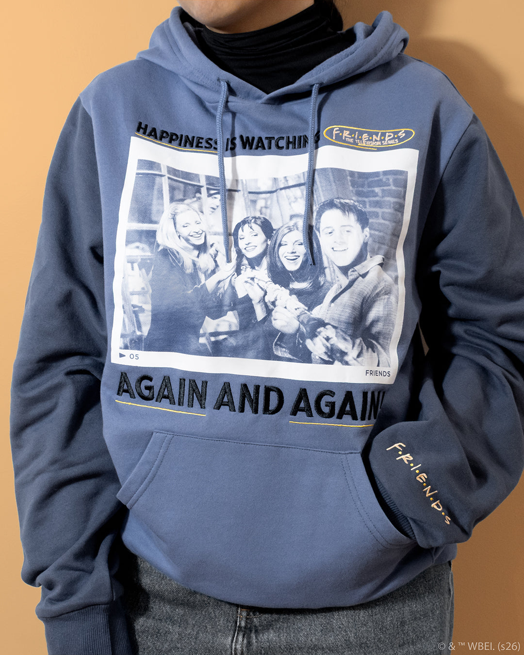 A person wearing a slate blue graphic hoodie with a large black-and-white photo print on the front and 'AGAIN AND AGAIN' text, paired with a black turtleneck and gray jeans against a warm beige background.