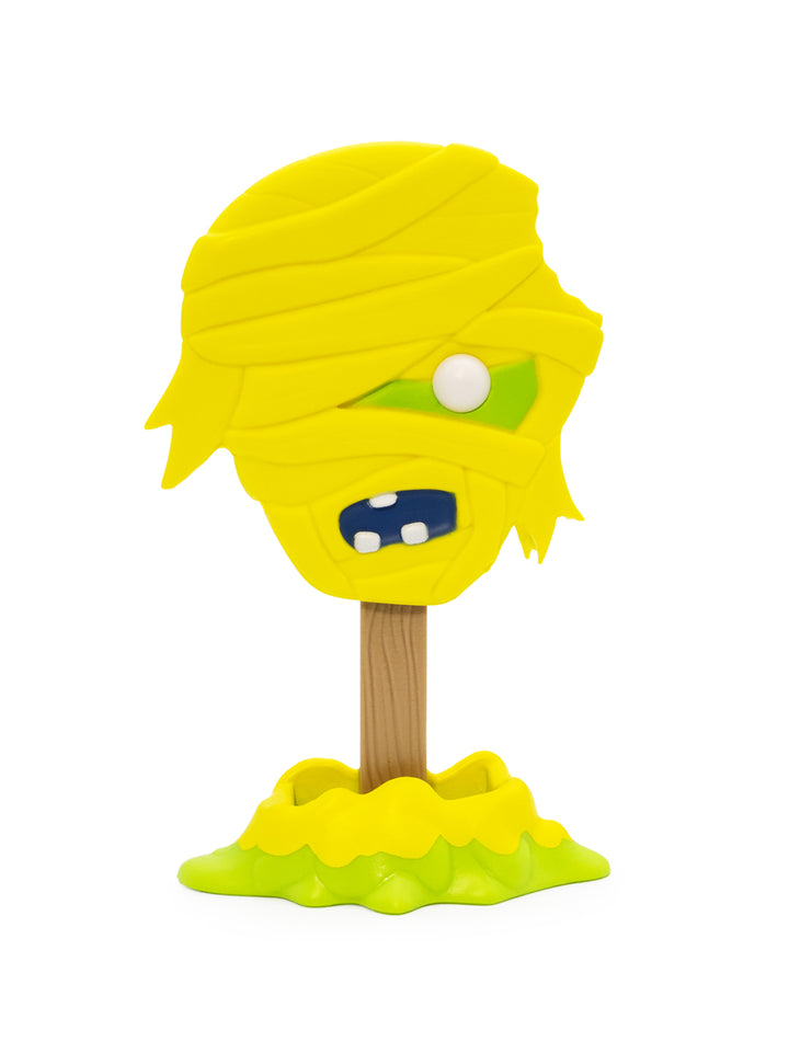 A yellow mummy-themed toy or decorative figure with a wrapped head, one visible white eye, dark blue mouth with white teeth, and a brown stick-like stem rising from a lime-green base, isolated on a white background.