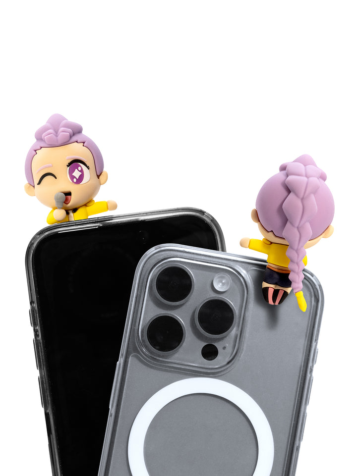 Two small purple-haired cartoon figurines wearing yellow tops are shown perched on the top edges of two smartphones, one black and one gray with a MagSafe ring, on a white background.
