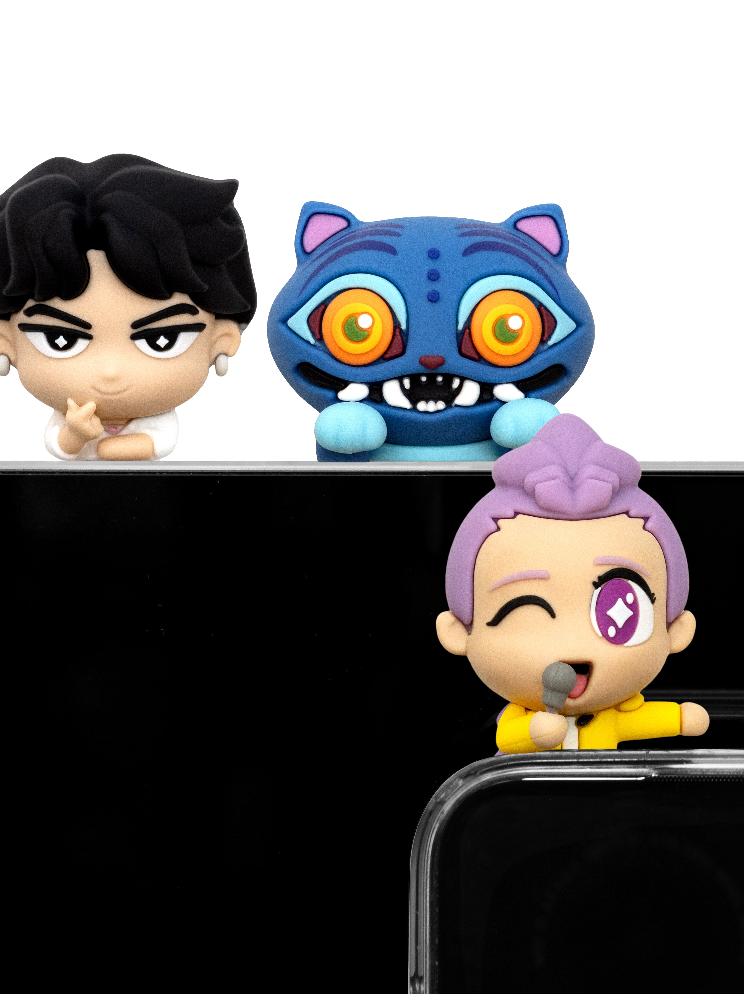 Three small colorful figurines or character toys arranged above a black surface on a white background: a black-haired character on the left, a blue cat-like character with large orange eyes in the center, and a purple-haired character with one eye closed on the lower right.