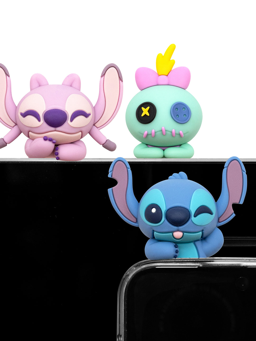 Three small character figurines displayed against a black background: a pink big-eared character on the left, a mint green character with a yellow hair and X-shaped eye on the right, and a blue character with large ears in the foreground. The foreground figure is partially cropped by a clear phone case.