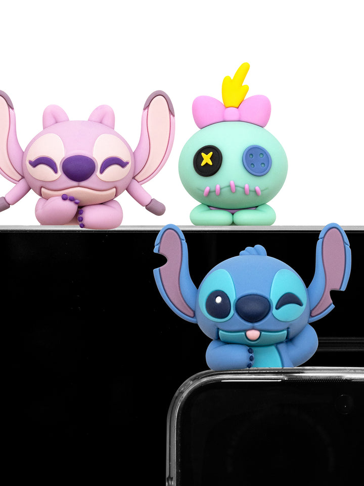 Three small character figurines displayed against a black background: a pink big-eared character on the left, a mint green character with a yellow hair and X-shaped eye on the right, and a blue character with large ears in the foreground. The foreground figure is partially cropped by a clear phone case.