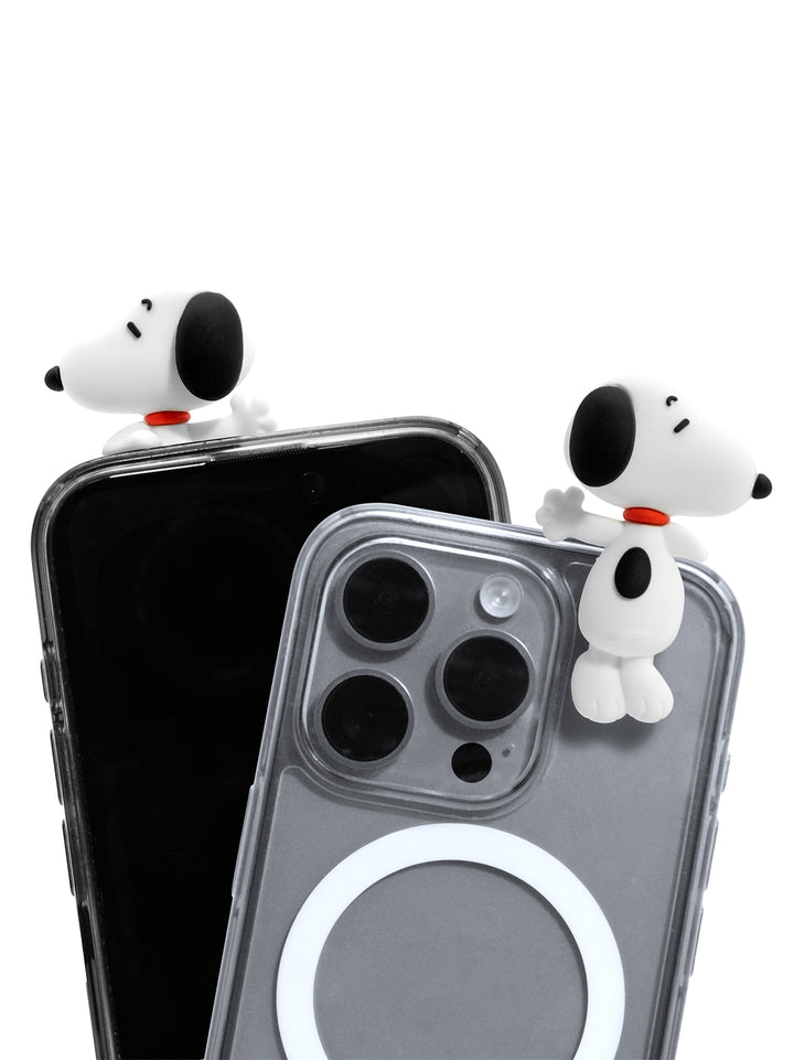 Two clear phone cases shown on smartphones against a white background, each with a small 3D Snoopy figure attached near the top edge. The front case is transparent with a white MagSafe ring visible, and the rear case is also clear.