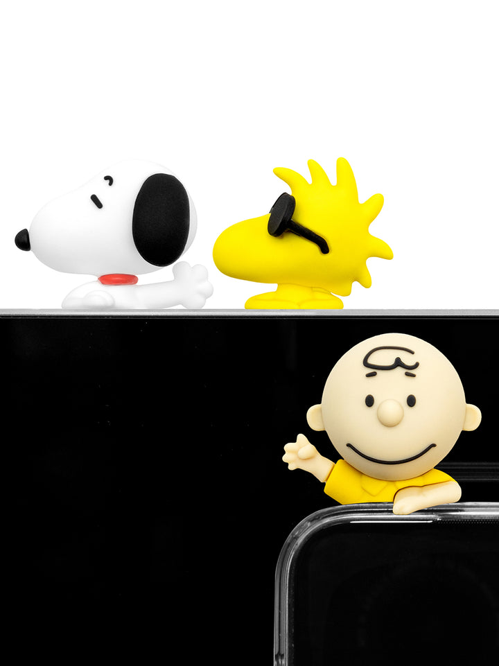 Three Peanuts character figurines, including Snoopy, Woodstock, and Charlie Brown, arranged on and around a black smartphone against a white background.