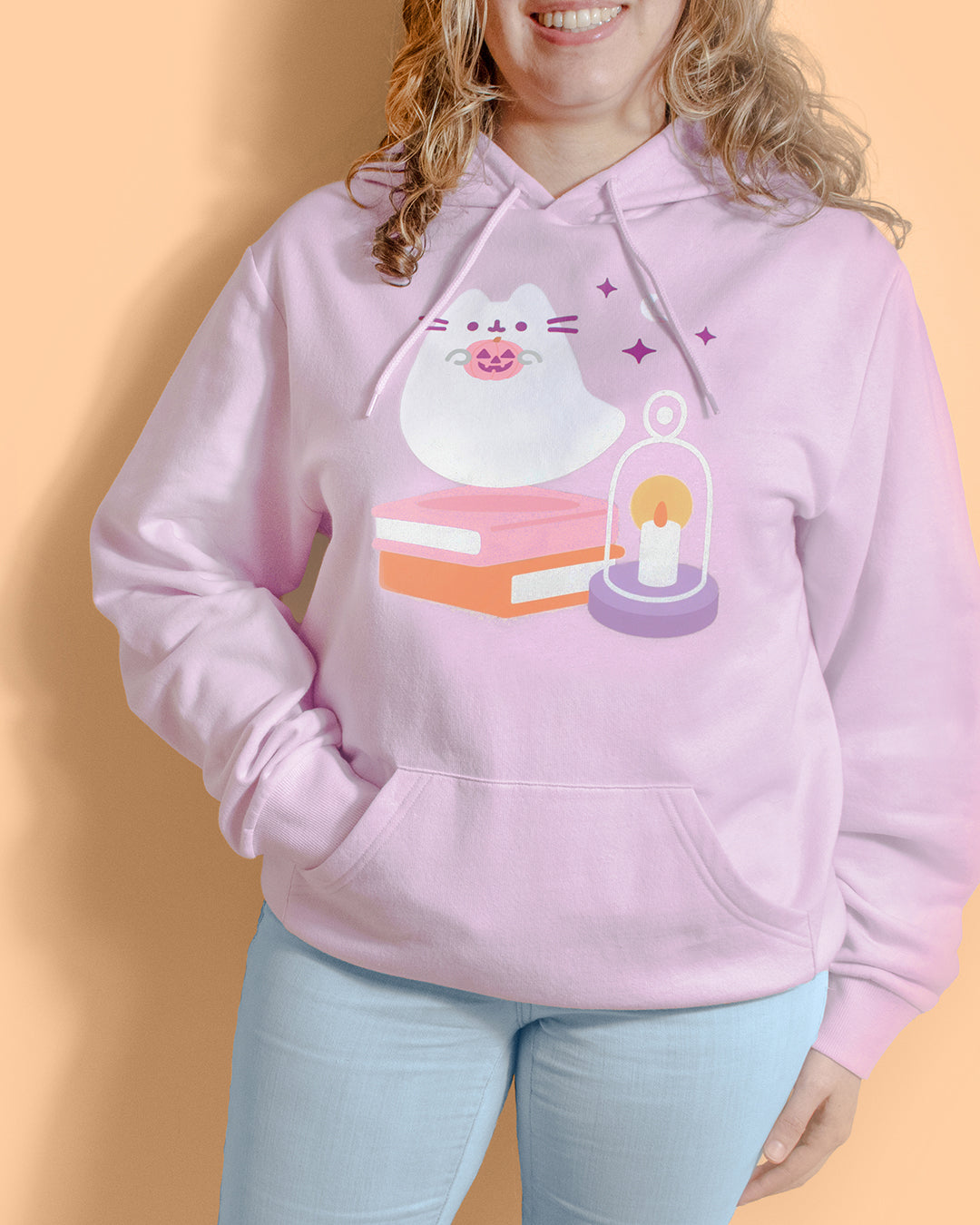 A woman wearing a light pink hoodie with a cute graphic of a white cat on a book next to a glowing candle under a glass dome, paired with light blue jeans against a solid peach background.