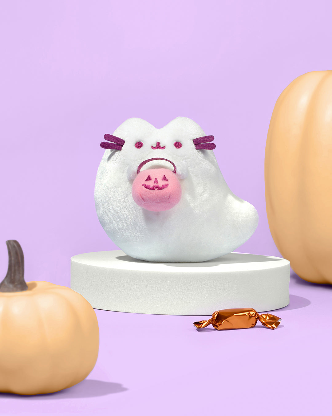 Ghost pusheen plush holding jack o lantern bucket. displayed in front of pumpkins and candy