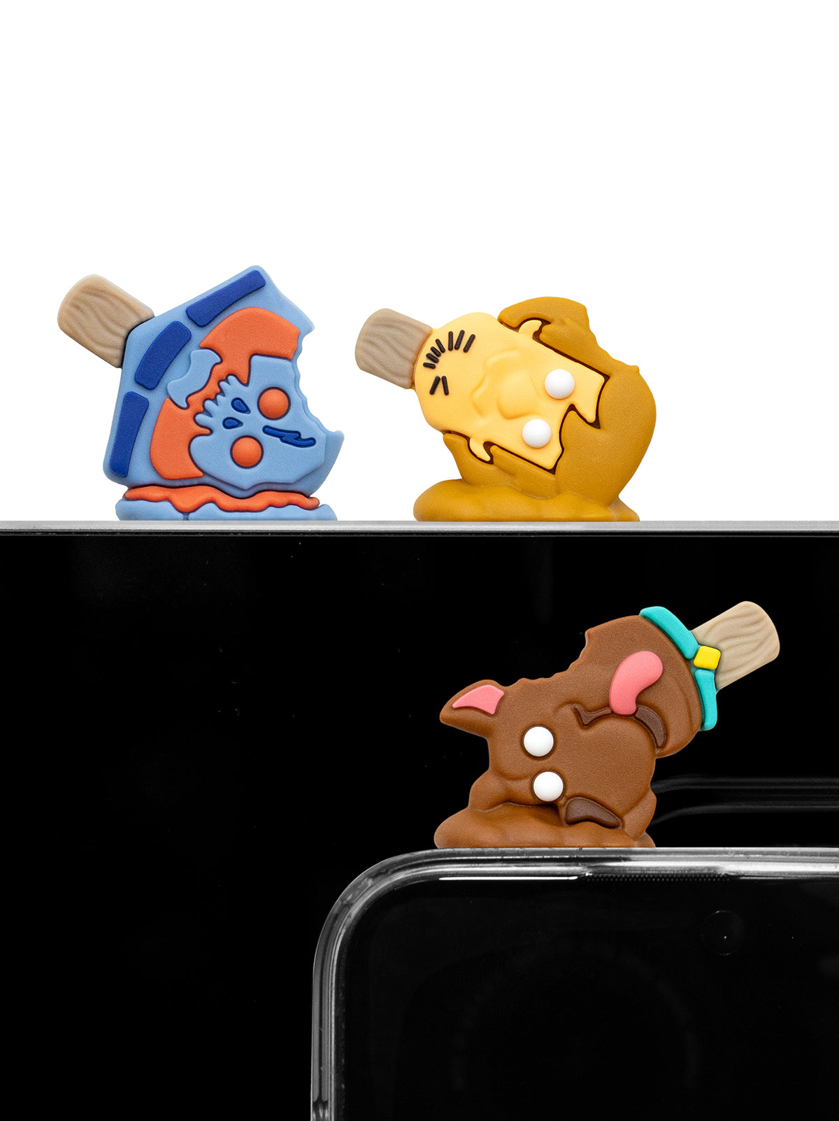 Three small novelty phone accessories shaped like cartoon characters are shown attached to the top and bottom edges of a smartphone. The top accessory is blue and orange, the middle accessory is tan and yellow, and the bottom accessory is brown with pink ears and a turquoise band.