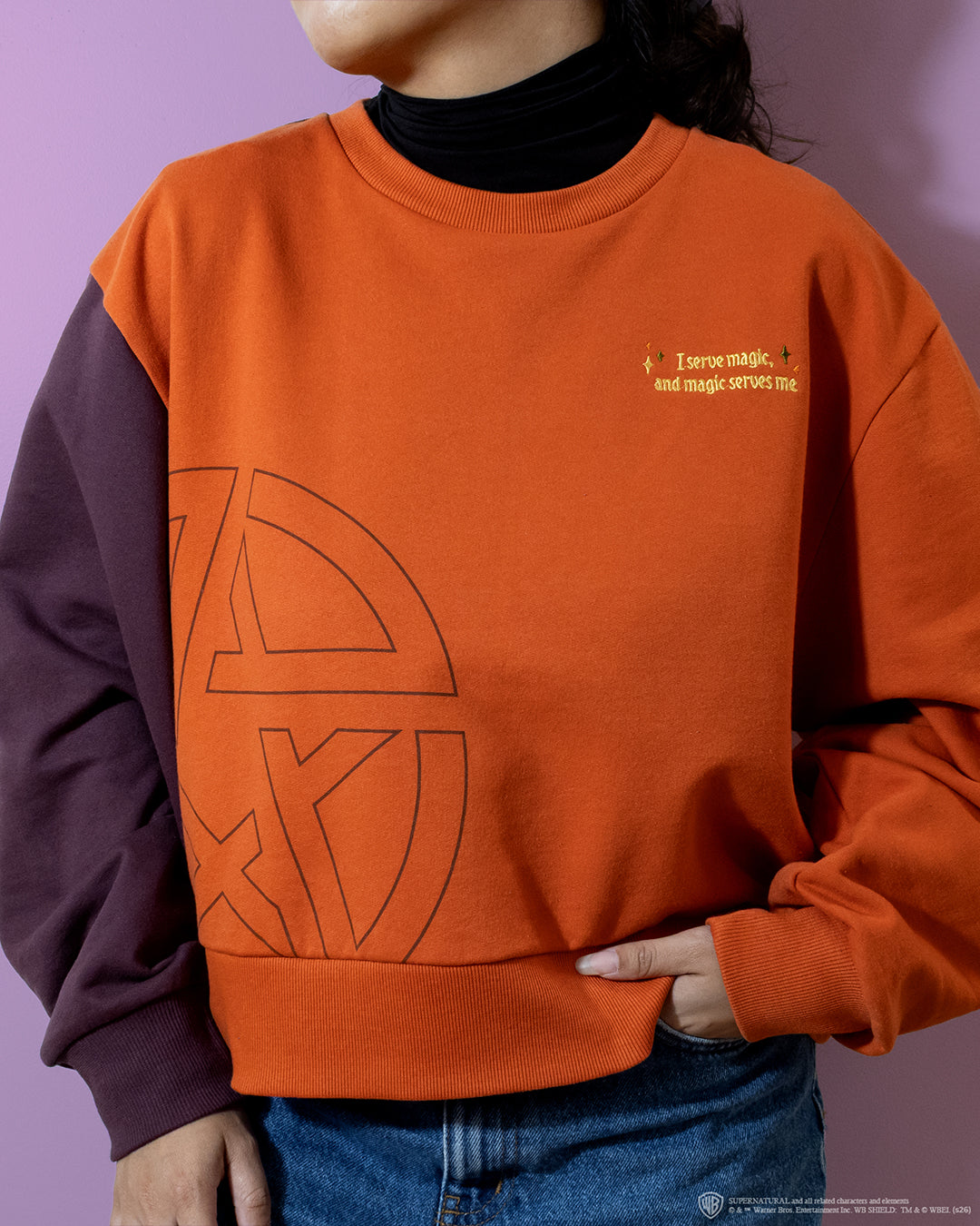 A person wearing a colorblock crewneck sweatshirt in burnt orange and dark plum, featuring a large geometric line-art graphic on the front and small yellow text on the left chest, paired with blue jeans and a black turtleneck.