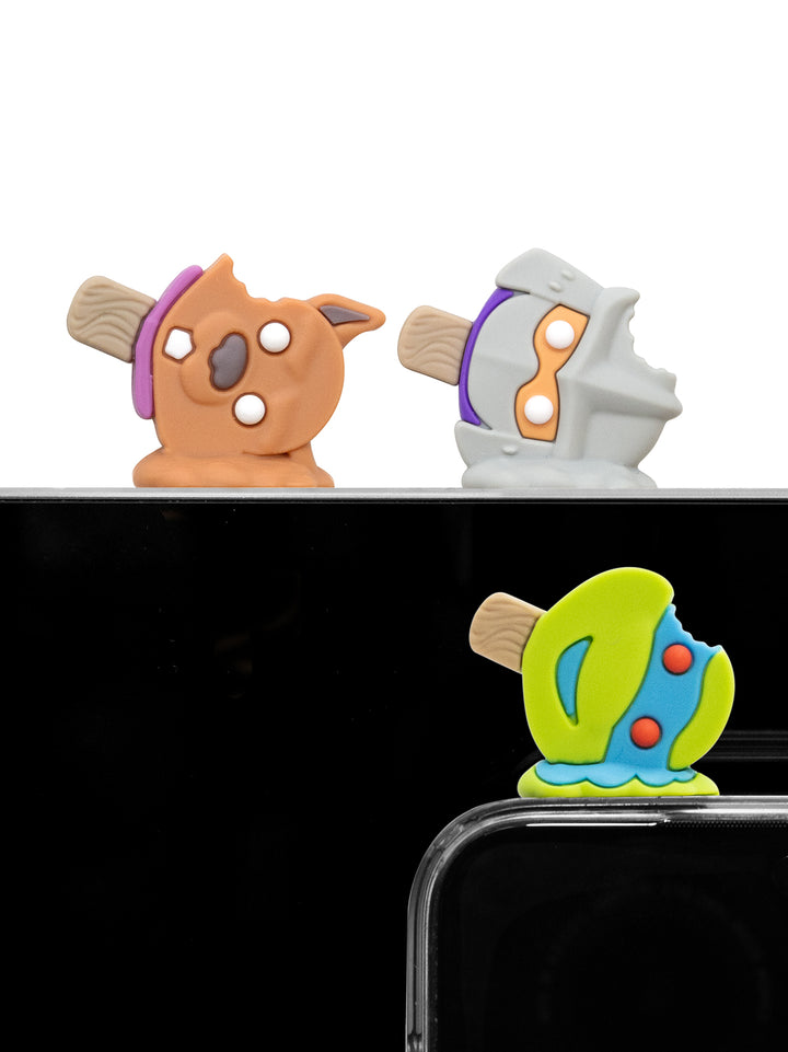 Three colorful novelty phone accessories shaped like cartoon characters, shown resting on a black surface with part of a smartphone visible at the bottom right.
