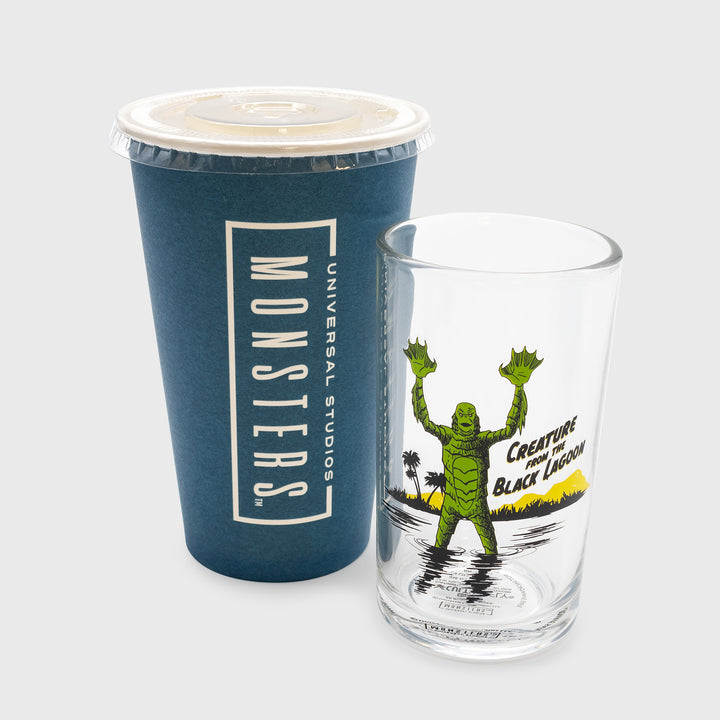 creature from the black lagoon glass. image on glass: sea monster raising out of water with hands up. palm trees, grass, and mountains in background. text: creature from the black lagoon. soda cup packaging next to it. text on cup packaging: universal studios monsters.