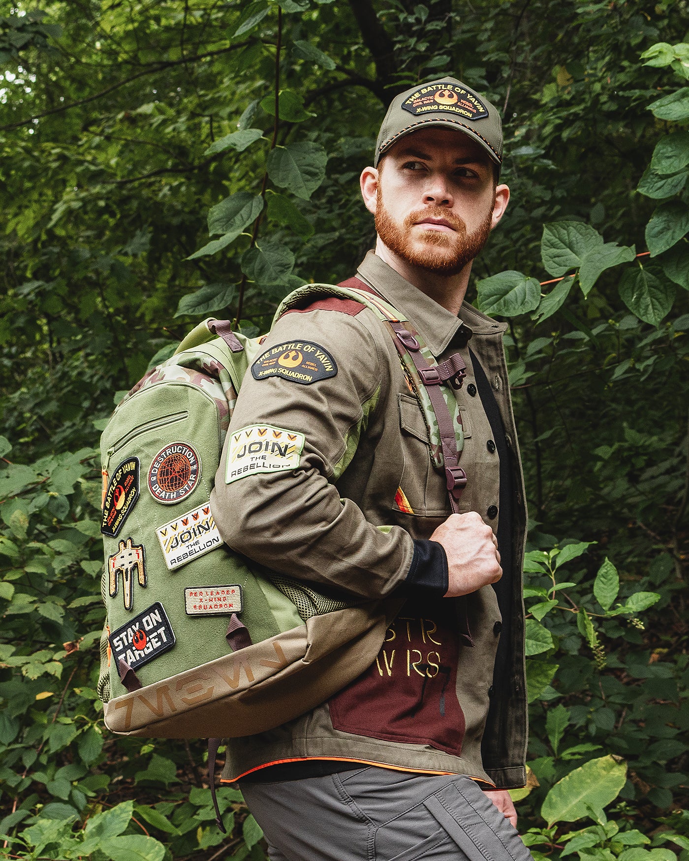 Man wearing a green jacket with patches and a backpack, standing in a forest.